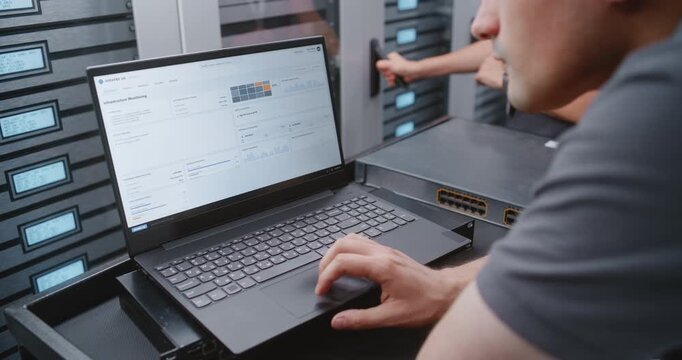 IT Administrator Uses Laptop Beside Network Switch to Navigate Server, Network and Security Logs, Comparing Software Metrics with Hardware Status Through LCD Displays in Data Center Infrastructure.