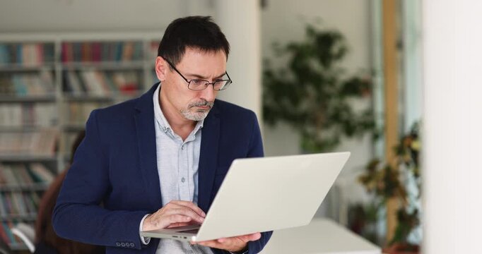 Focused middle aged businessman professor or teacher researching academic information on laptop in library analyzing data planning lectures or lessons preparing materials for personal or online course