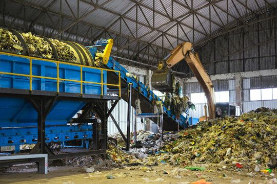 Heavy excavator loading piles of municipal solid waste onto an industrial trommel screen conveyor system inside