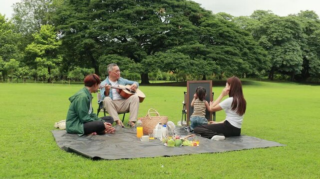GrandFather playing acoustic guitar for child during picnic. Asian family enjoy outdoor picnic with food and drinks in park sunny day, outdoor activitie with daughter father, mother grandfather