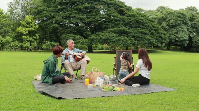 GrandFather playing acoustic guitar for child during picnic. Asian family enjoy outdoor picnic with food and drinks in park sunny day, outdoor activitie with daughter father, mother grandfather