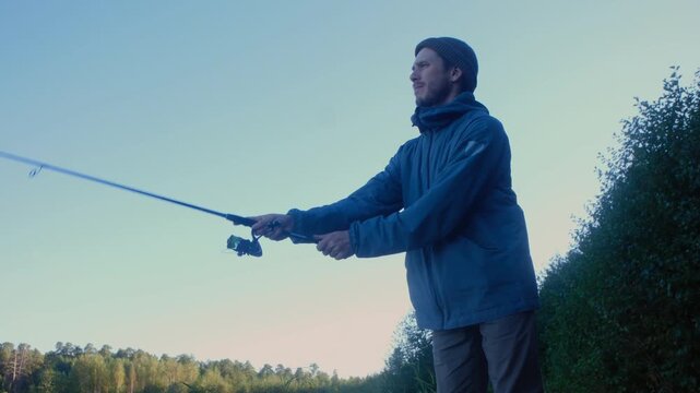 Young man in blue jacket and beanie hat casting lure with spinning rod while fishing on lake, with beautiful calm forest in background and clear blue sky. Fisherman casting spinning rod into lake
