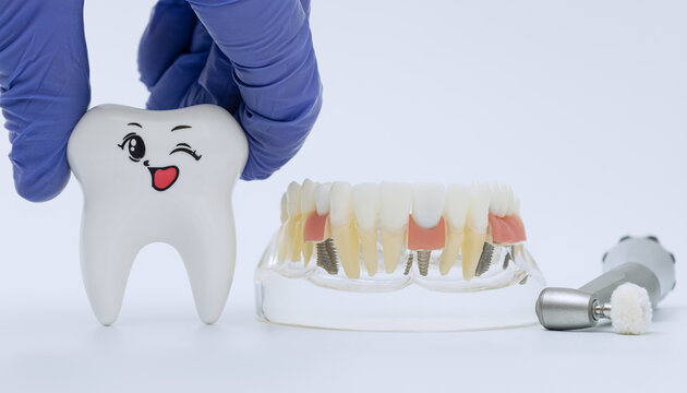 Overcoming dental fear concept with doctor hand holding a funny smiling tooth character near a clear acrylic prosthesis model and medical handpiece.