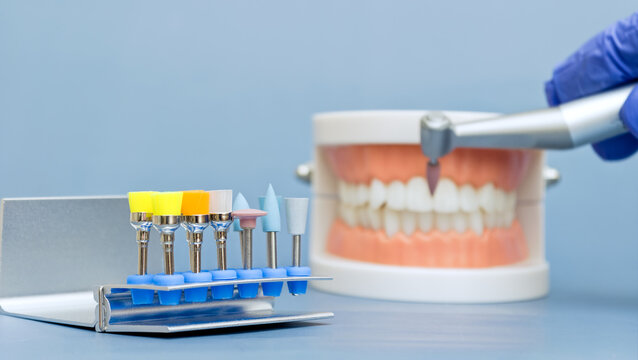 Set of colorful dental polishing burs in sharp focus. Blurred dentist hand holding drill handpiece treating jaw model in background.