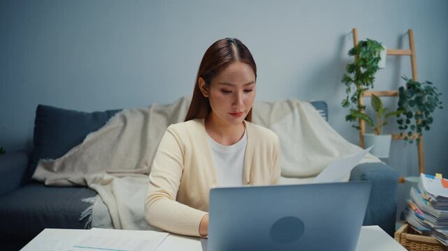 Young Asian woman reviews financial documents, bills and expenses on laptop at home. Personal finance budget planning, Tax invoice paperwork review, Household savings and expense management concept.