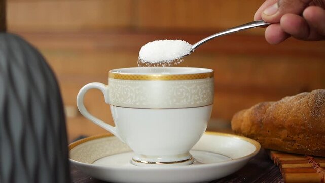 Close up shot of a person adding white granulated sugar to a steaming cup of tea or coffee, slow motion footage