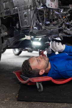 Auto mechanic inspecting vehicle undercarriage with work light while lying on creeper, focused repair service in garage