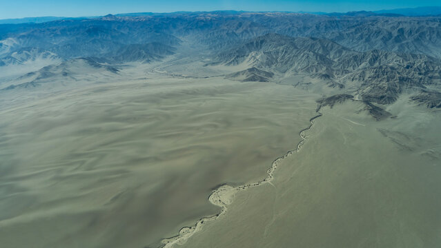 The Nazca Plateau. An aerial view, from an airplane. The bed of a dried‑up river winds through the sands and dunes of the desert. Mountains devoid of vegetation. A blue sky. Peru.