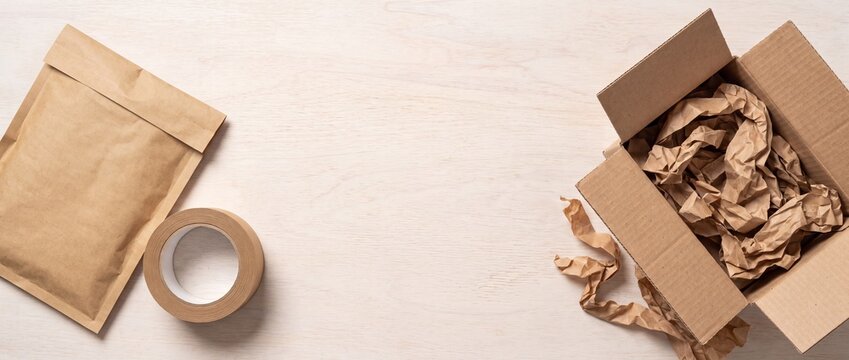 Open cardboard box with packing paper envelope and tape roll on a light wooden surface with crumpled brown packing sealed paper mailing sits next sealed eco friendly stationery recyclable parcel