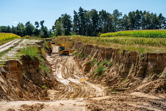 Wide angle shot of a deep sandy excavation pit with a yellow tracked excavator working inside a ravine, flanked by steep sandy cliffs, green agricultural fields, and a forest tree line under a clear b