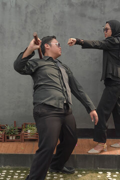 Asian Muslim woman in hijab throwing a punch at man holding wooden stick.