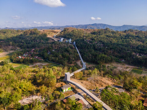 Aerial Scenery of Covered Conveyor Belt System with Checkpoint Buildings and a Service Road Crossing Hilly Terrain in Indonesia. Industrial Infrastructure Extends Across the Green Hillside. 