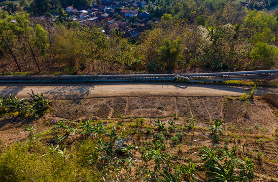 Aerial View of a Covered Conveyor Belt System with Checkpoint Buildings and a Service Road Crossing Hilly Terrain Near a Residential Area in Indonesia