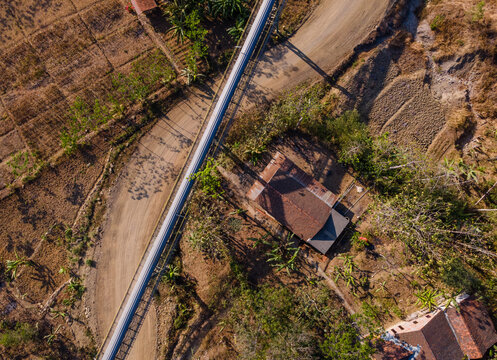 Aerial View of a Covered Conveyor Belt System with Checkpoint Buildings and a Service Road Crossing Hilly Terrain Near a Residential Area in Indonesia