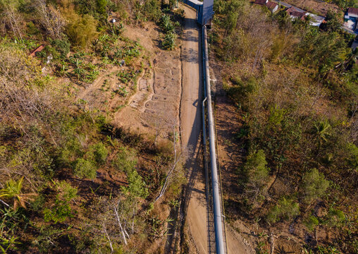 Aerial View of a Covered Conveyor Belt System with Checkpoint Buildings and a Service Road Crossing Hilly Terrain Near a Residential Area in Indonesia