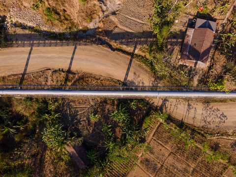 Aerial View of a Covered Conveyor Belt System with Checkpoint Buildings and a Service Road Crossing Hilly Terrain Near a Residential Area in Indonesia