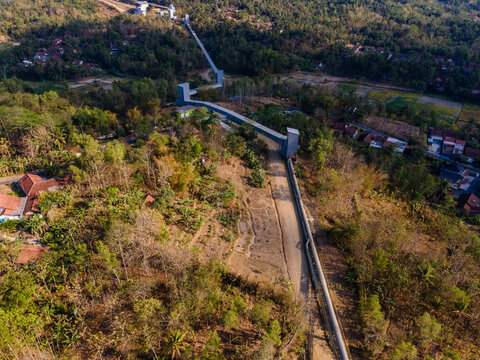Aerial View of a Covered Conveyor Belt System with Checkpoint Buildings and a Service Road Crossing Hilly Terrain Near a Residential Area in Indonesia