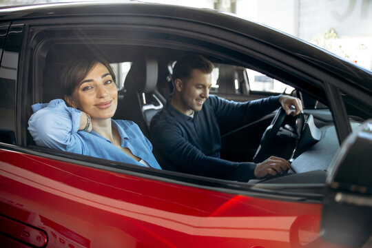 Young couple looking for a new car at a dealership, a woman smiling at the camera sitting inside a modern red vehicle