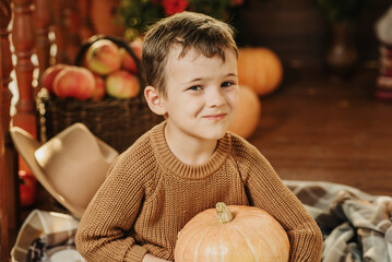 Portrait of a Caucasian boy in a sweater holding a pumpkin on the porch of his house in the autumn season. © Anastasia