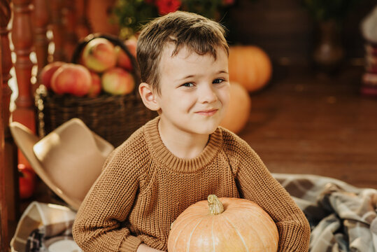 Portrait of a Caucasian boy in a sweater holding a pumpkin on the porch of his house in the autumn season.