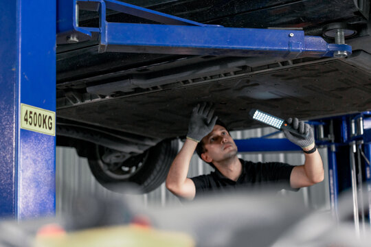 Mechanic inspecting car underbody with work light under vehicle on lift in modern garage