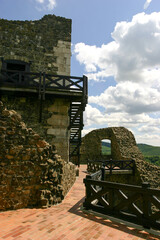 A view of the ruins of a stone castle with wooden walkways and stairs, set against a backdrop of green hills and a cloudy blue sky. © Nick