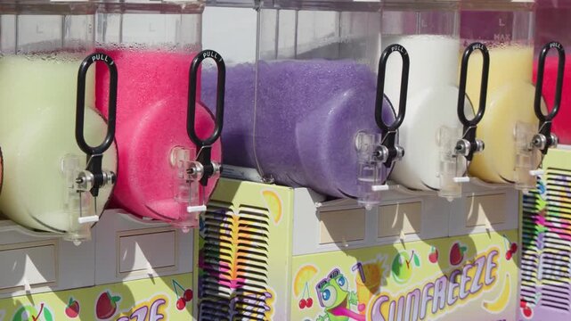 Slushie machine with colorful frozen drinks rotating and mixing at outdoor carnival event.