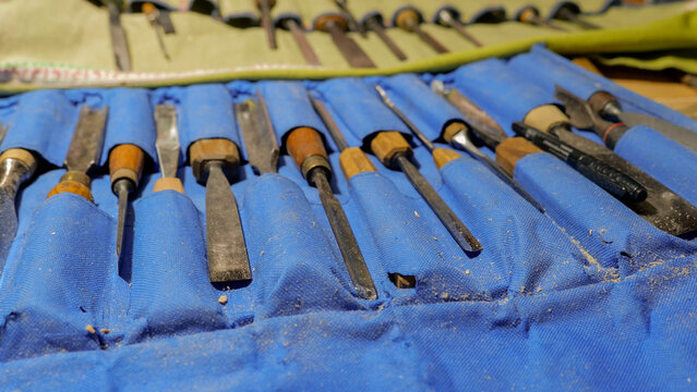 Set of woodcarving chisels and gouges arranged in blue fabric roll with wood shavings, worn wooden handles, metal blades, and shallow depth of field in workshop light