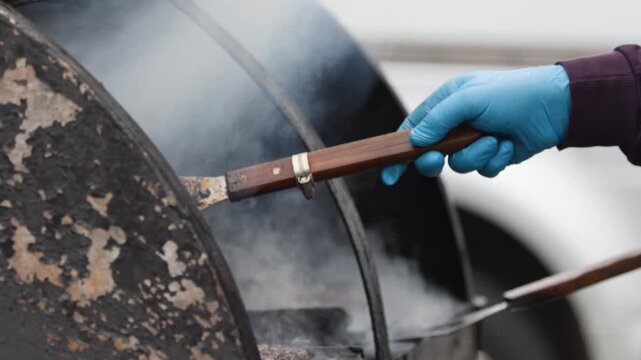 Close up of hand in blue glove using tongs to flip burger patties on outdoor grill with smoke rising.