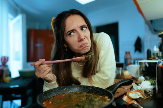 Disgusted Young Woman Holding Spoonful Of Ruined Food In Kitchen. Frustrated lady looking away with dislike while holding unappetizing cooked meal sample