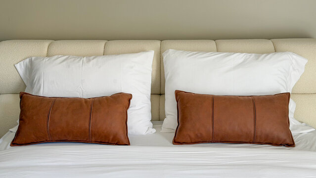Modern cozy bed with white bedding and brown decorative pillows, neutral bedroom interior with soft upholstered headboard and minimalist hotel style.