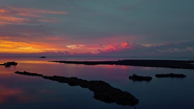 KALOCHORI, GREECE: Cinematic aerial video flying forward over a tranquil lagoon at sunset, featuring a small wooden boat, flying birds, and colorful clouds reflecting on the calm water.