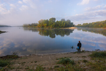 A person stands on the calm riverbank, watching the quiet morning fog rise over a lush, forested island reflected in water © Александр Арендарь
