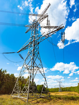 Power lines stand tall against blue sky. Electricity transmission towers are in a field with trees and clouds in the background. It is daytime and sunny.