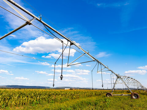 Center pivot irrigation system over agricultural corn field. A metal sprinkler structure extends across a green crop field under a bright blue sky with clouds.