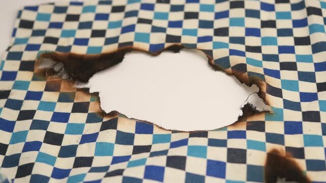 Burned hole in blue and white checkered fabric with charred edges showing white background