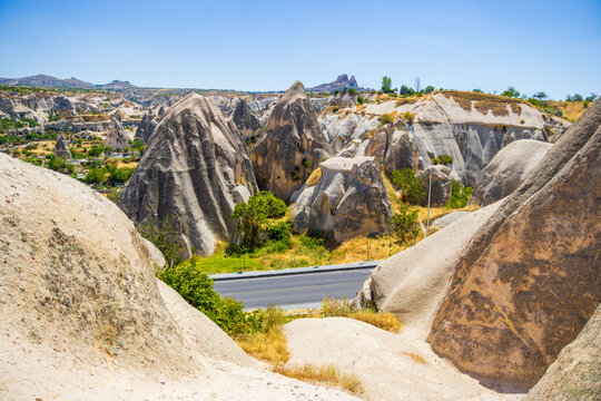 Rugged volcanic rock formations surrounding a winding paved road beneath a clear blue sky in a sunlit Cappadocia landscape of Turkey