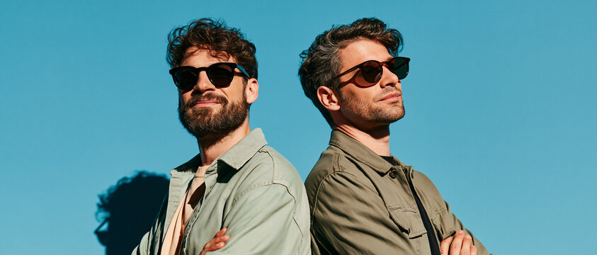 Two stylish men wearing sunglasses and casual jackets stand back-to-back against a vibrant blue sky. One man has a beard and a slight smile, while the other looks away with a confident expression.