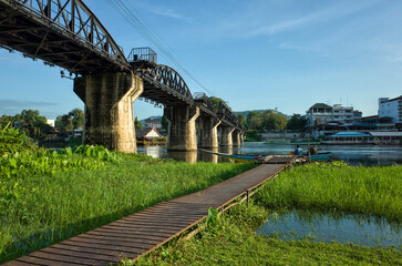 Steel truss bridge over the River Kwai stands above calm water and boats, framed by green banks and bright, clear sky © art_of_line