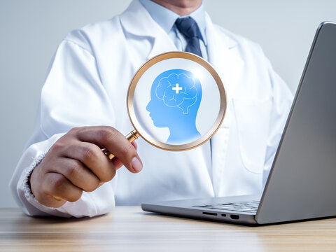 Clinician holds magnifying glass over brain and head symbol beside laptop. Digital cognitive assessment, mental health screening, neurological evaluation, and modern clinical diagnostics concept.