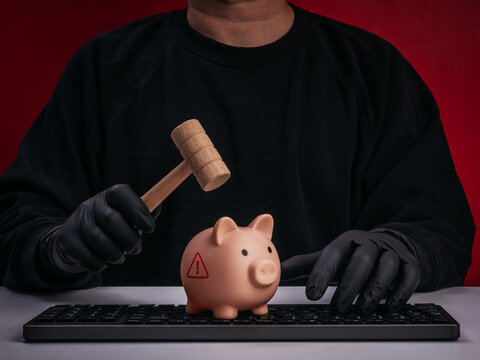 Gloved scammer or hacker raises a mallet above warning-marked piggy bank on computer keyboard. Online financial fraud, threatened savings, cybercrime, scams, theft, and consumer protection concept.