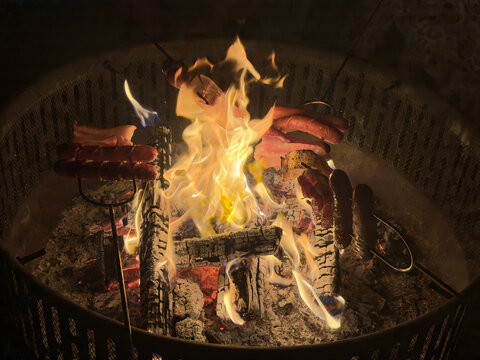 Delicious hot dogs and bacon roasting on metal skewers over a vibrant campfire in a backyard fire pit at night, capturing a cozy and rustic outdoor cooking experience.