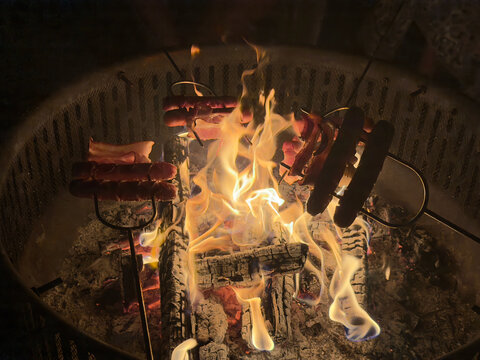 Delicious hot dogs and bacon roasting on metal skewers over a vibrant campfire in a backyard fire pit at night, capturing a cozy and rustic outdoor cooking experience.