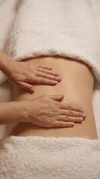 Hands perform gentle abdominal lymphatic drainage massage across a woman's stomach between white towels, using smooth manual strokes on the exposed midsection in a clean spa treatment setting.