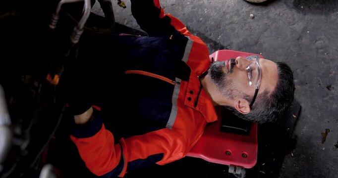 Adult male mechanic lies on rolling creeper beneath electric vehicle, moving gloved hands while inspecting undercarriage components during preventive maintenance inside automotive service workshop.