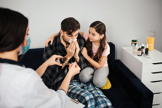 Sick young amn sitting on sofa and sneezing. His daughter sitting beside. She comfort him. Female doctor pouring some syrup into spoon.
