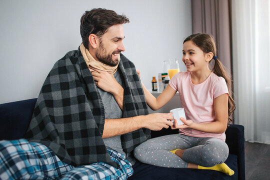 Positive picture of young man sitting together with his daughter. They smile to each othr. Guy is sick. They hold white cup together.