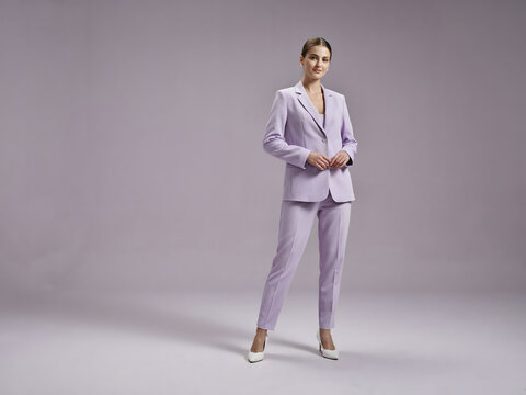 A woman with dark hair wearing a light purple blazer and matching trousers with white heels standing against a soft light purple background