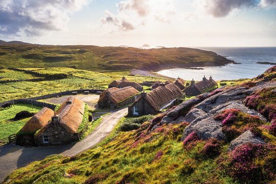Gearrannan Blackhouse Village with thatched stone cottages, heather and coastal bay at golden hour, Isle of Lewis, Outer Hebrides, Scotland