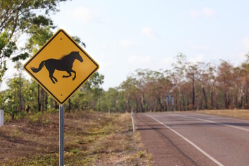 Yellow horse crossing road sign cautioning drivers for safety on a quiet outback highway with bushland and trees under a clear sky with lots of copy space for animal warning themes in Australia © BJP7images